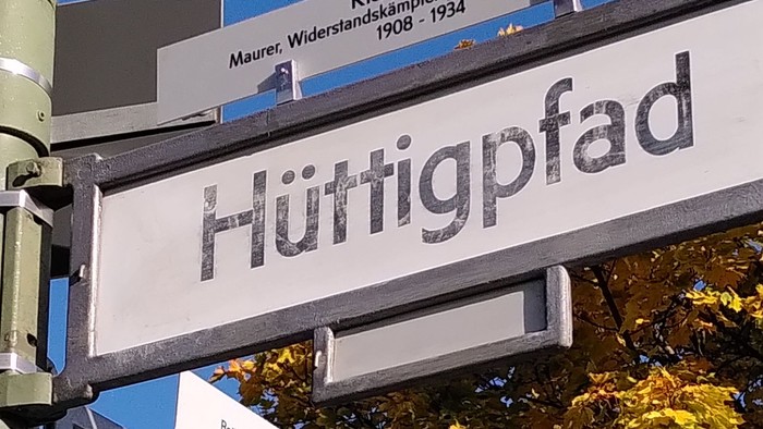 Schilder mit den Straßennamen Hüttigpfad und Emmy-Zehden-Weg sowie zusätzliche Gedenktafeln in einem städtischen Gebiet.