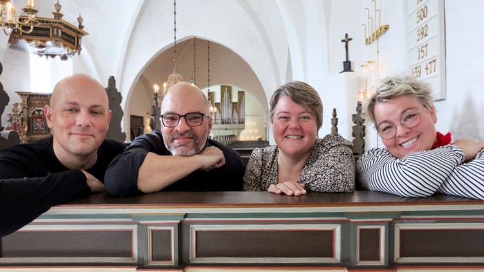 Fire personer poser glade sammen i et prægtigt kirkeinteriør med træbænke og buer.