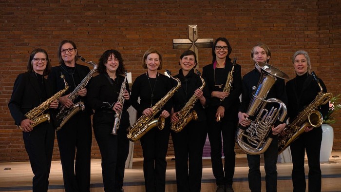 Eine Gruppe von Musiker:innen, die mit verschiedenen Blechblasinstrumenten in einer kirchenähnlichen Umgebung posieren.