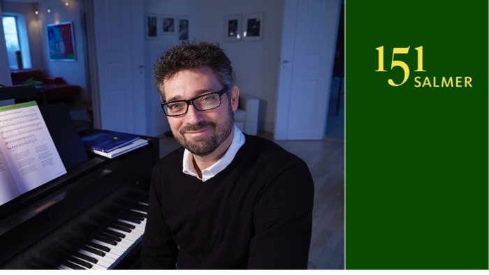 En mand med briller står ved siden af et klaver med noder, iført en sweater og smilende.