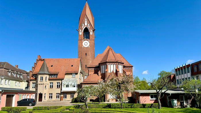 Bild zeigt eine Kirche mit hohem Turm und Uhr, umgeben von einem gepflegten Garten.