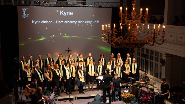 Ein Chor singt auf einer Bühne mit großem Kronleuchter und Projektion 