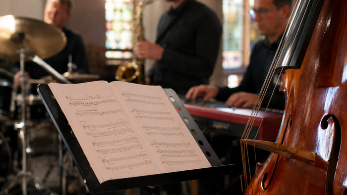 Nahaufnahme eines Notenständers mit aufgeschlagenen Notenblättern. Im Hintergrund sind unscharf Mitglieder einer Jazzband in einer Kirche zu erkennen: ein Schlagzeuger, ein Saxophonist, ein Pianist und ein Kontrabass. Das warme Licht und die farbigen Kirc