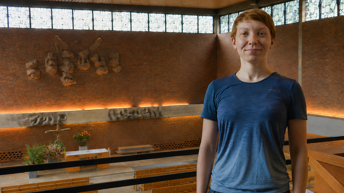 Eine Person steht auf einer Empore in einer modernen Kirche und blickt in die Kamera. Im Hintergrund öffnet sich der Blick in den Kirchenraum mit Altar, Kreuz, Blumen und einer großflächigen Backsteinwand mit künstlerischer Gestaltung. Das warme Licht unt