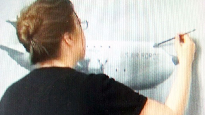 Person, die ein großes Wandgemälde eines Flugzeugs mit der Aufschrift „U.S. Air Force“ an einer Wand malt.