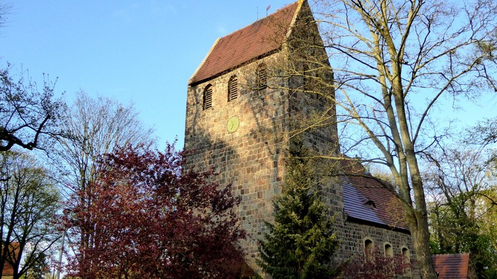 Alte Kirche mit hohem Turm und rotem Dach, umgeben von Bäumen und Büschen