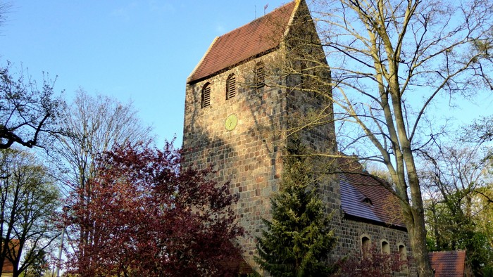 Alte Kirche mit hohem Turm und rotem Dach, umgeben von Bäumen und Büschen