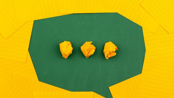 Zerknautschte orangefarbene Papierkugeln in einer grünen Sprechblase auf einem knallgelben Hintergrund.