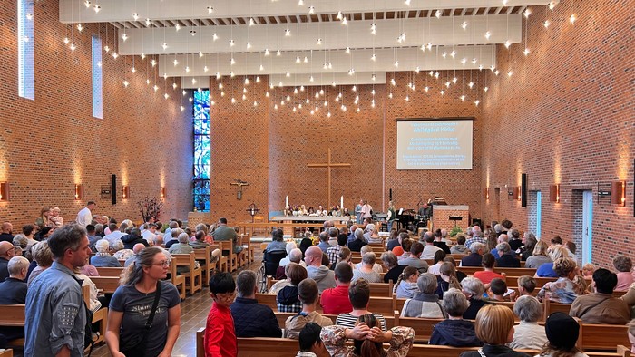 En overfyldt kirkes indre med deltagere, der sidder og står, og deltager i en gudstjeneste med en stor skærm og alter.