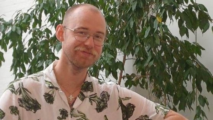 En skaldet mand med briller iført en skjorte med bladmønster læner sig op ad et bord ved siden af en grøn plante.