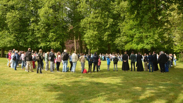 Eine große Gruppe von Menschen versammelte sich im Freien auf einer grasbewachsenen Fläche, umgeben von Bäumen, und schien zu warten oder etwas zu beobachten.