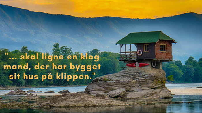 En hytte på en klippe ved en elv med bjerg i baggrunden.