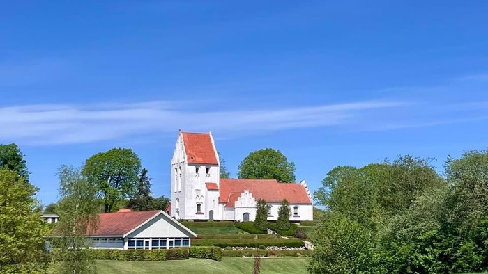 En fredelig kirke med røde tage står midt i frodig grønne omgivelser under en klar blå himmel.