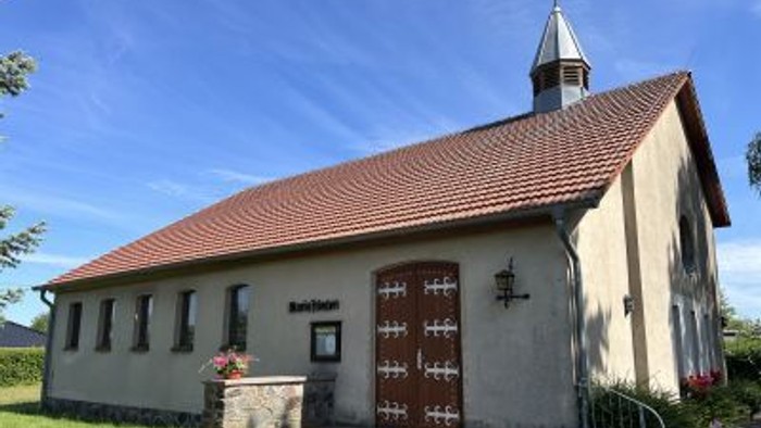 Idyllische Kirche mit rotem Ziegeldach, hölzernen Doppeltüren und kleinem Turm in grüner Umgebung.
