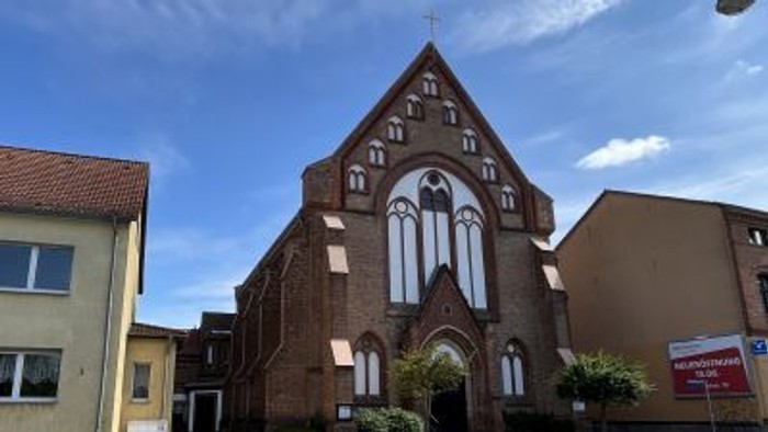 Eine Backsteinkirche mit hohen Spitzbogenfenstern und einem steilen Satteldach steht markant an einer Straßenecke.
