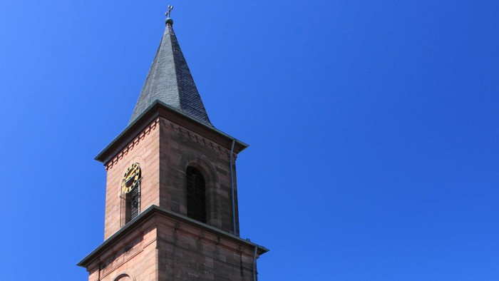 Kirche mit hohem Uhrturm und Spitzhelm vor klarem blauen Himmel.