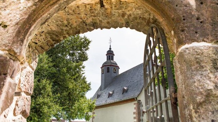 Ein steinerner Torbogen rahlt einen Uhrturm und ein Gebäude mit steilem Dach ein.
