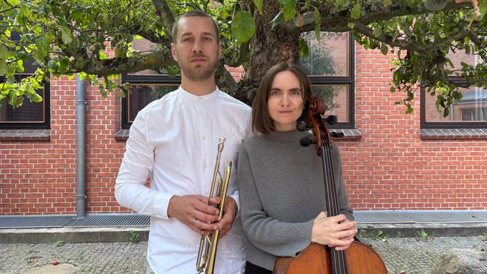 To musikere, der holder en trompet og en cello, står udendørs nær en murstensbygning med træer.