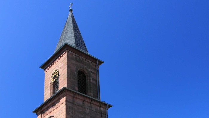 Kirche mit hohem Uhrturm und Spitzhelm vor klarem blauen Himmel.
