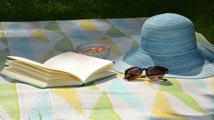 Ein Buch, eine Sonnenbrille und ein Strohhut auf einem bunten Picknicktuch im Freien
