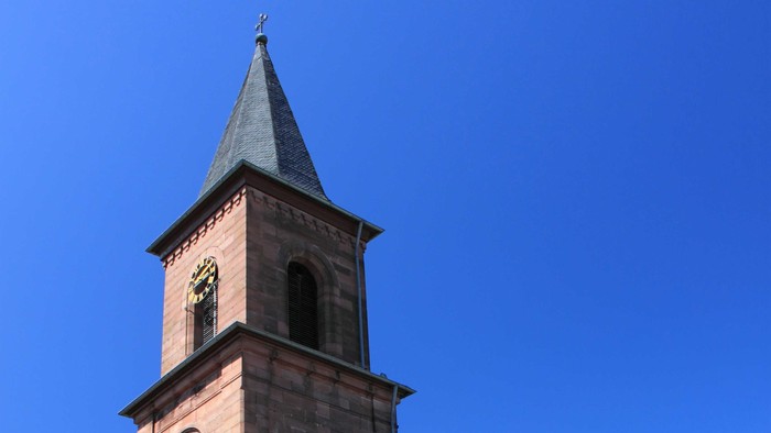 Kirche mit hohem Uhrturm und Spitzhelm vor klarem blauen Himmel.