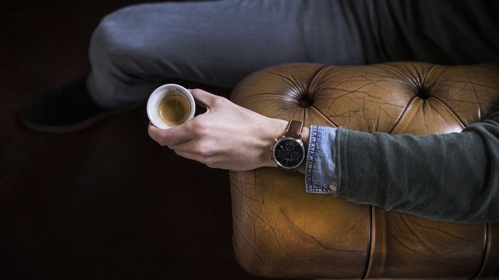 En person sidder på en brun læderstol og holder en kop kaffe.