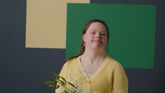 En kvinde i gul trøje står foran en mørk blå vegg med grønne og gule rektangler og en blomst.