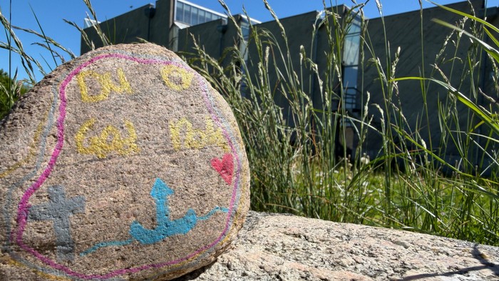 Farverig malet sten med motiver placeret på en ru overflade omgivet af højt græs og en kirke i baggrunden.