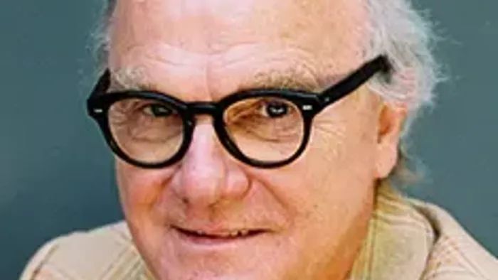 Lächelnder älterer Mann mit runder Brille, kariertem Sakko und rotem Hemd.
