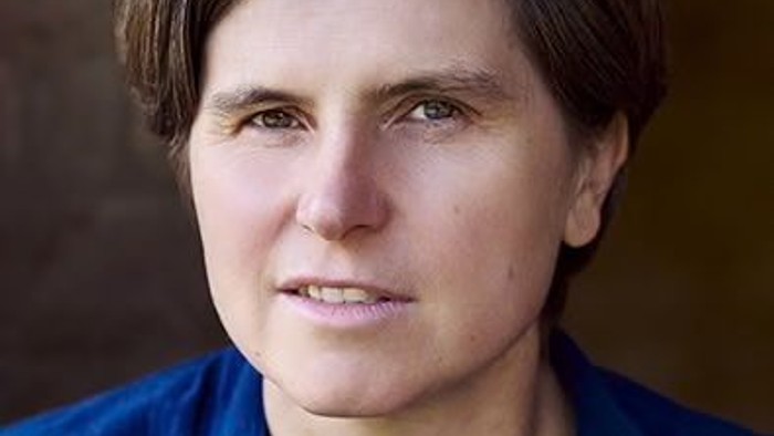Porträt einer Person in blauem Hemd vor neutralem Hintergrund.