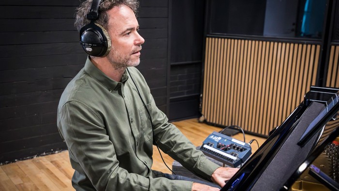 En mand med hovedtelefoner optager eller producerer musik i et lydisoleret studie ved hjælp af et keyboard og et mixerbord.