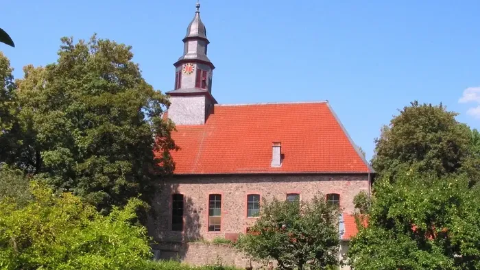 Idyllische Steinkirche mit rotem Dach und Glockenturm, umgeben von üppigem Grün unter klarem Himmel.