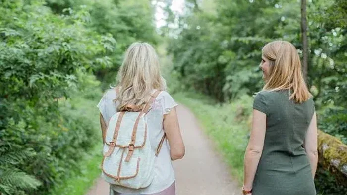 Zwei Frauen gehen gemeinsam einen üppigen, grünen Waldweg entlang, eine trägt einen Rucksack.
