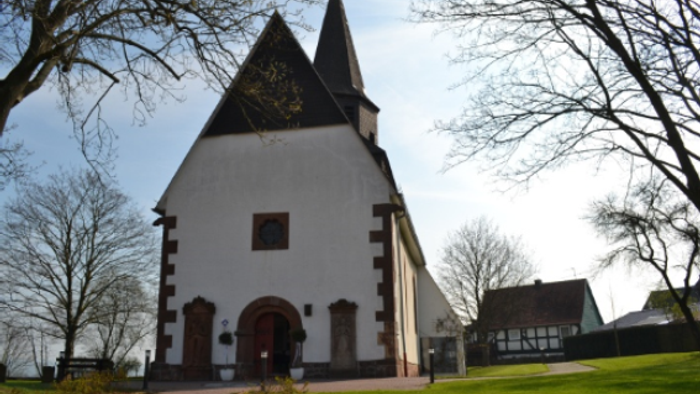 Idyllische weiße Kirche mit steilem Satteldach und dunklem Turm, umgeben von kahlen Winterbäumen und einer grasbewachsenen Fläche.