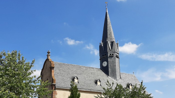 Kirche mit hohem Turm und Uhr, umgeben von Bäumen und Blumen.