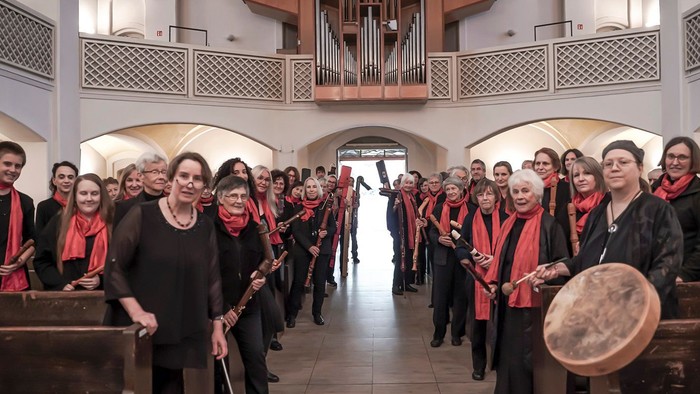 Gruppe von Menschen in roten Schals und schwarzer Kleidung, einige halten Musikinstrumente, stehend in einer großen Halle mit einer Orgel im Hintergrund.