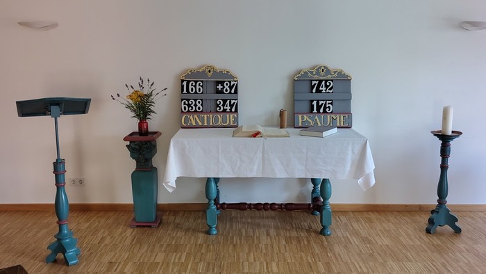 Minimalistischer Altarraum mit Kerzen, Tafeln und einfachen Holzstühlen in einem hellen Raum.