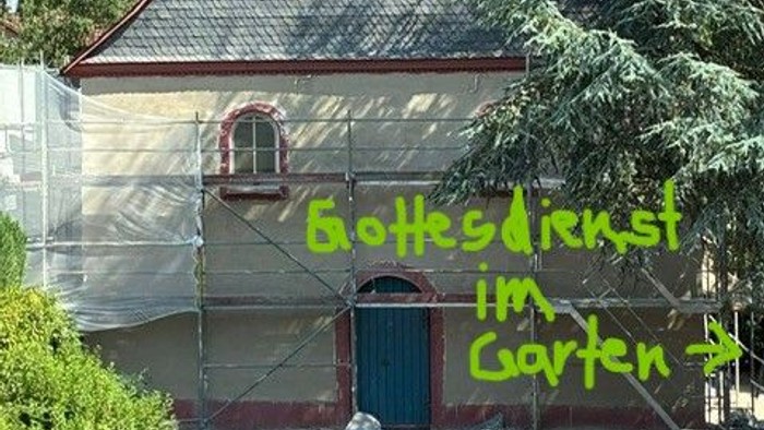Eine Kirche im Umbau mit Gerüsten, die mit dem Schild „Gottesdienst im Garten“ auf Freiluftgottesdienste hinweist.