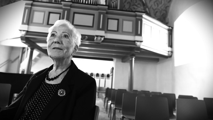 Ældre kvinde i formelt tøj betragter eftertænksomt i en kirke- eller kapelscene.