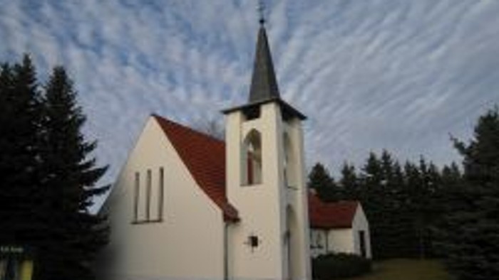 Idyllische weiße Kirche mit rotem Dach und Turm, eingebettet in eine ruhige, bewaldete Landschaft.