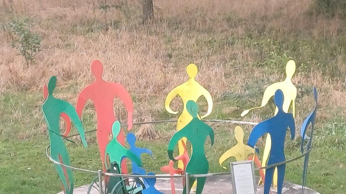 Flächige bunte Körper aus Metall im Kreis, verschiedene Größen, einer mit Rolli. Auf einer Wiese. Im Hintergrund Bäume und vertrocknetes Gras.