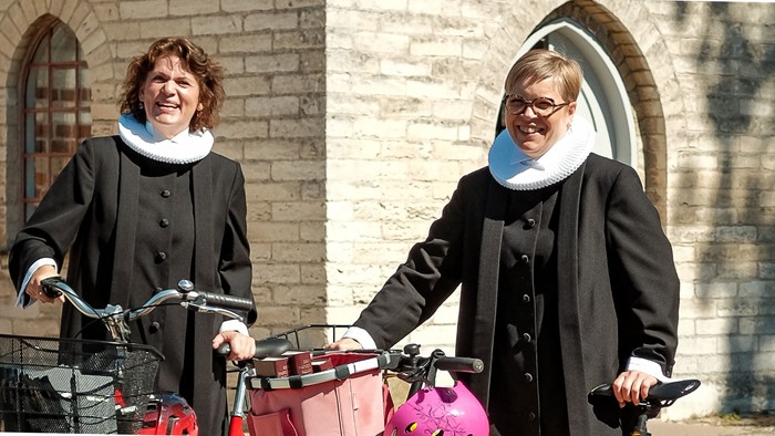 To kvinder i formelt tøj poser ved siden af cykler med indkøbsposer, smilende udendørs nær en murstensbygning.