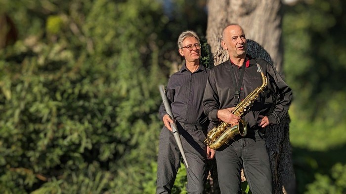 Zwei Männer in einer Outdoor-Umgebung, einer hält ein Saxophon und der andere eine Gitarre, sie stehen in der Nähe von Grünpflanzen.
