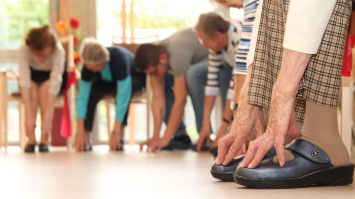 Eine Gruppe älterer Menschen nimmt an einem Fitnesskurs teil, alle bücken sich nach vorne.