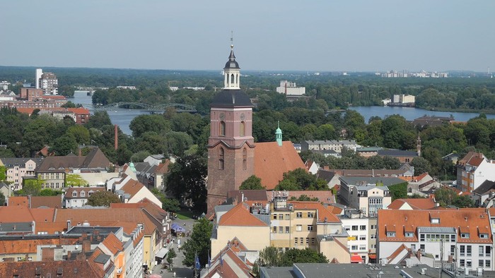 Luftaufnahme einer historischen Stadt mit einem markanten Kirchturm und einem sich schlängelnden Fluss durch grüne Umgebung.