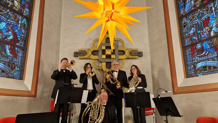 Ein Blechbläserensemble spielt in einer Kirche, die mit farbenfrohen Buntglasfenstern und einer goldenen Sternendekoration geschmückt ist.