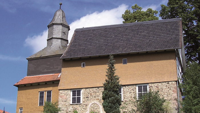 Historische Steinkirche mit hohem Glockenturm und efeubewachsenen Mauern vor klarem Himmel.