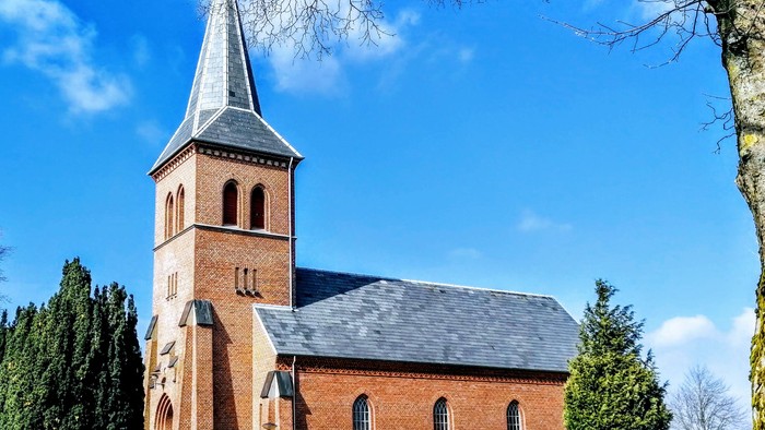 Hyggelig rødstenskirke med højt spir mod en klar blå himmel og frodig grønne omgivelser.