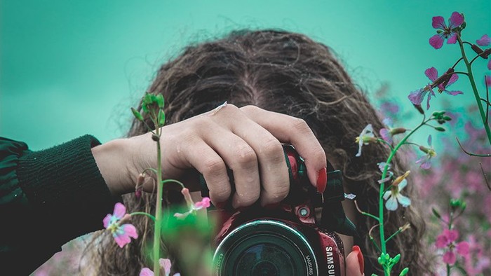 Frau fotografiert mit großer Kamera durch Blumen hindurch.