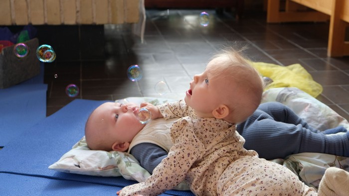 To babyer leger på måtter med bobler, hvor den ene rækker ud efter svævende sæbebobler, mens den anden ligger i nærheden.